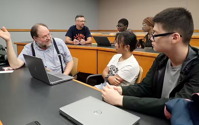 Professor leading a small-group discussion with students using laptops in a classroom.