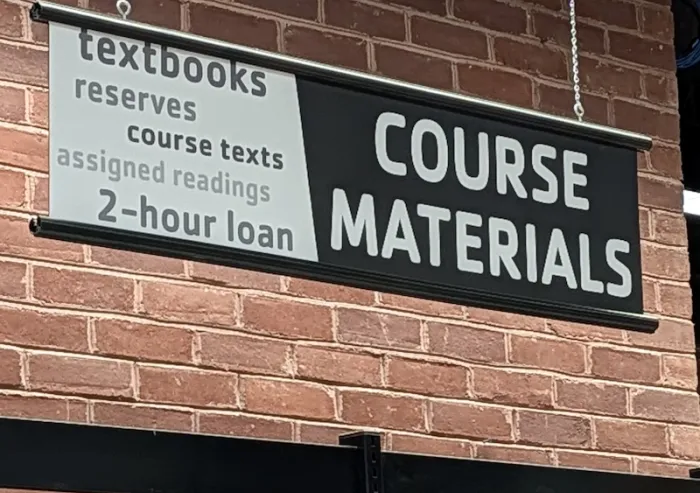 A black and white sign with Course Materials printed on it, hanging in front of a red brick wall.