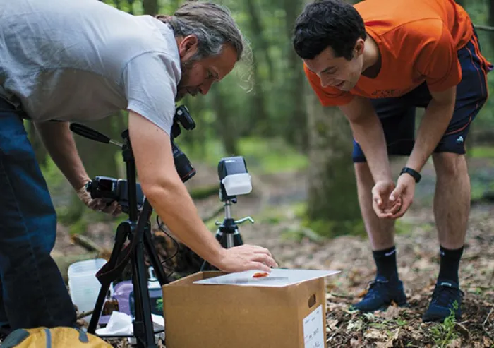 Two people work outdoors in a wooded area, handling equipment beside a cardboard box.