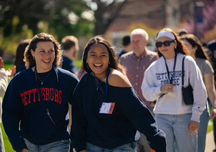 Two students walk outdoors with a larger group on a tree-lined campus path.