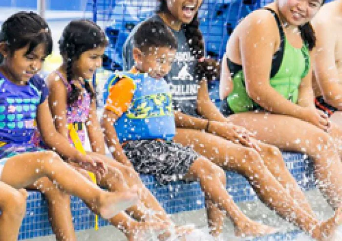 A group of students and young children sit at the edge of a swimming pool with their feet splashing in the water.