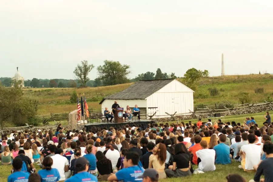 photo of students seated in field in front of speaker on stage with battlefields in background