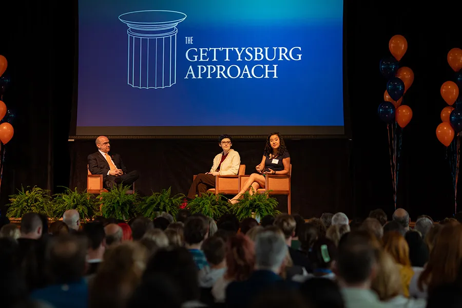 President Iuliano and students sit on stage in front of crowd with The Gettysburg Approach logo on screen in background