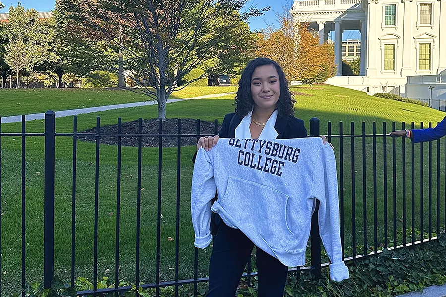 female student poses in front of White house holding up Gettysburg College sweatshirt