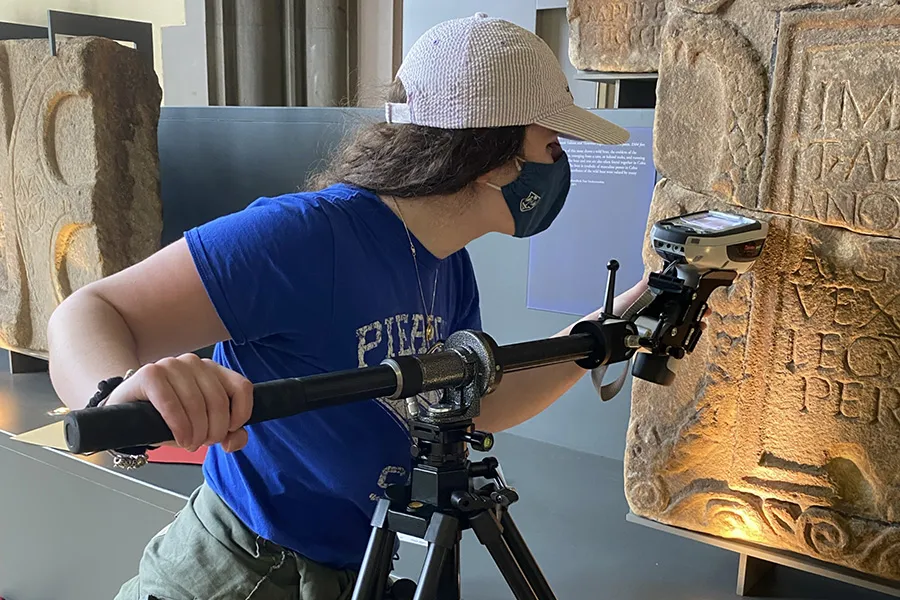 masked student in ballcap uses technology to scan stone tablet
