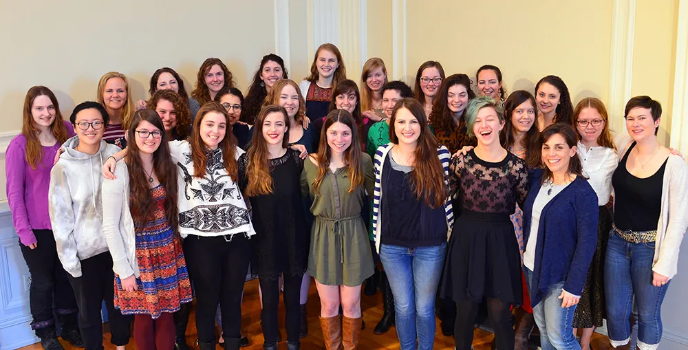 A group of women from the Audeamus choir poses together indoors, smiling at the camera in a formal room.