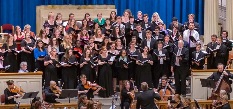 A large choir sings with an accompanying string orchestra in a church or concert hall, led by a conductor.