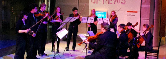 A small string ensemble performs indoors, with violinists and violists gathered around music stands under colorful stage lighting.
