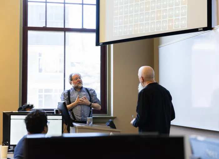 prof and visiting speaker talk at front of classroom