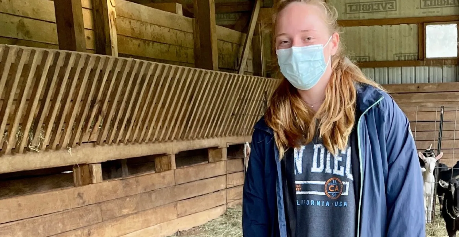 masked female student stands in barn with goats in backround