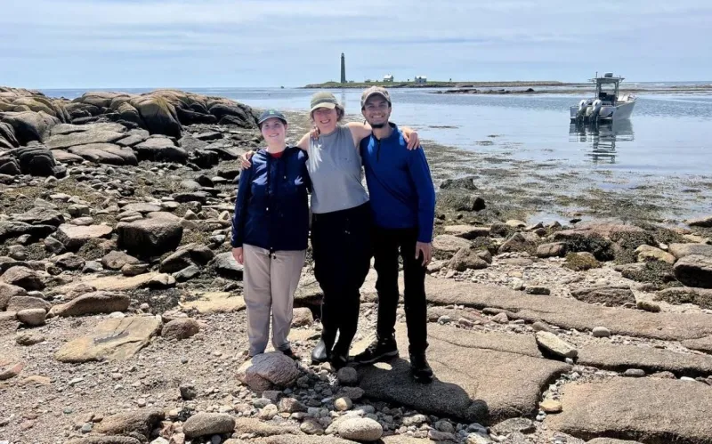 students pose on rocky beach with ocean, boat, and lighthouse in background